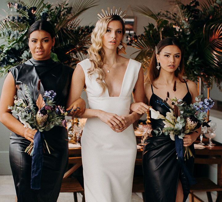 Bride in a fitted wedding dress with plunging neckline and a gold crown flanked by her bridesmaids in leather dresses 