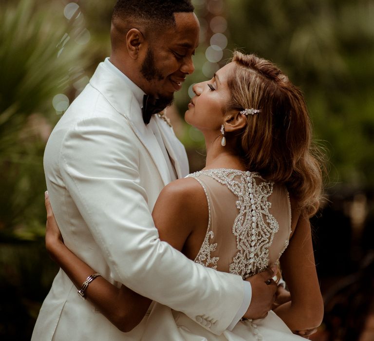 Groom in a white jacket embracing his bride with side swept wavy hair in a Justin Alexander dress with embellished back detail and buttons 