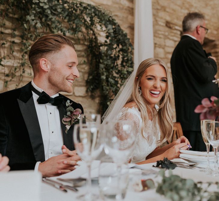 Laughing bride in white lace Augusta Jones top sitting with groom in black Hugo Boss suit and bow tie at top table for stone barn Caswell House wedding