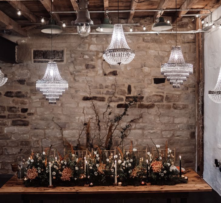 Intimate wedding reception with wooden table, chandelier light installation and foliage and flower table flowers