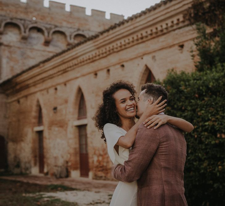 Groom in a brown check suit kissing his smiley bride with naturally curly hair 