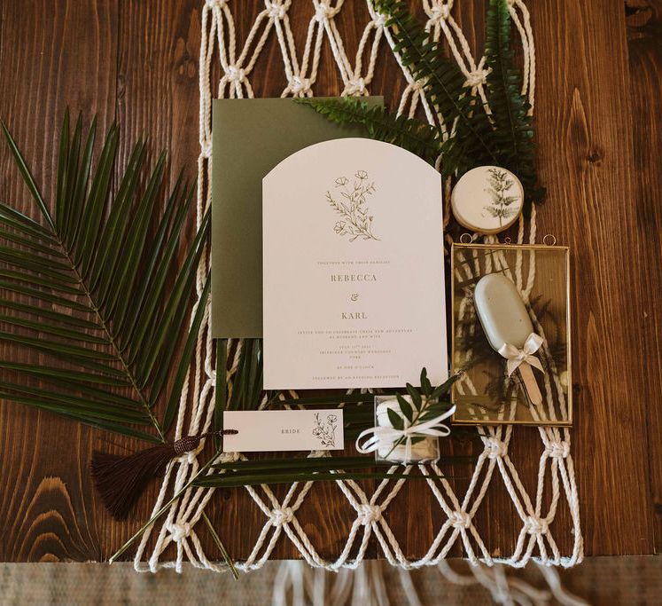 Wedding stationery lies upon macrame backdrop on wooden table with green foliage