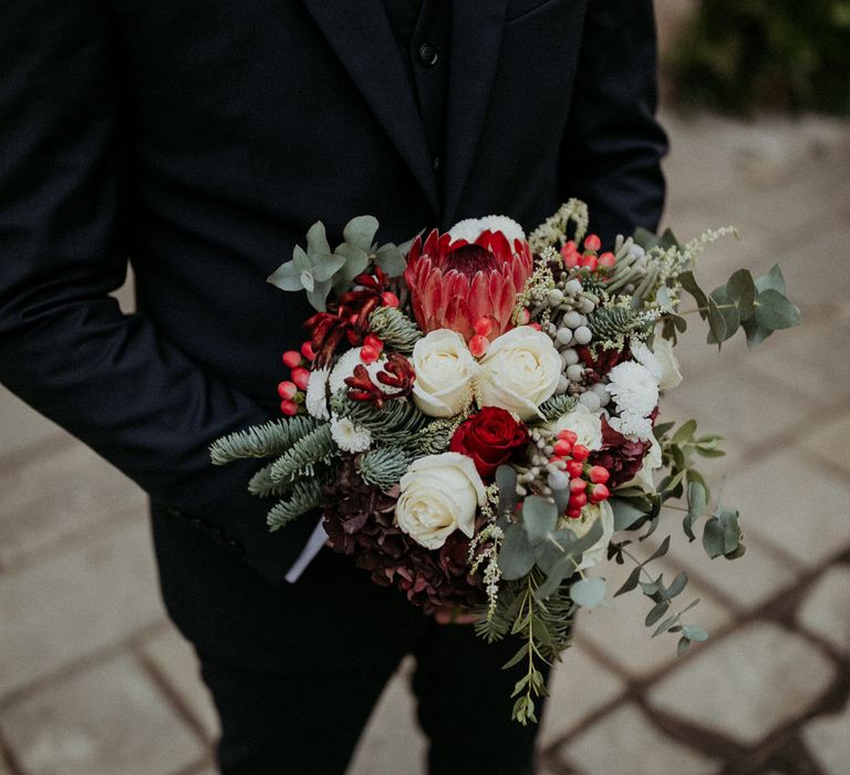 The groom holding a bouquet of red and white roses, eucalyptus and pine leaves, berries and seasonal blooms