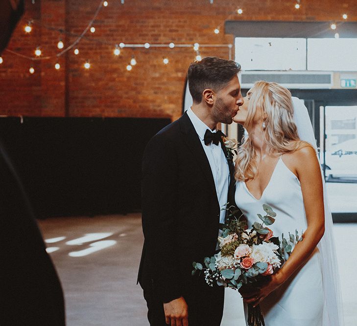 Bride in a slip wedding dress holding a peach and green bouquet kissing her husband 