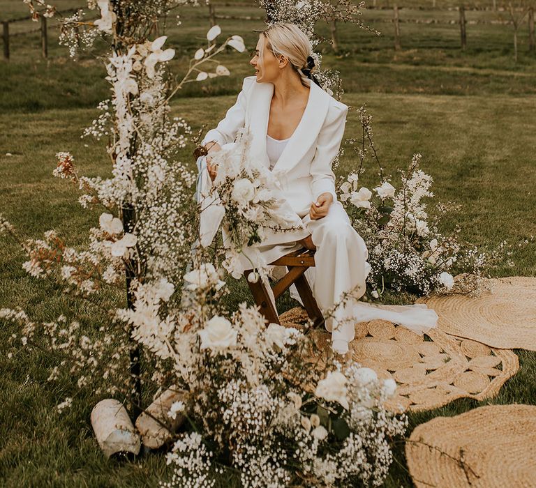 Bride in a white suit sitting on a wooden stool under a white flower arch 