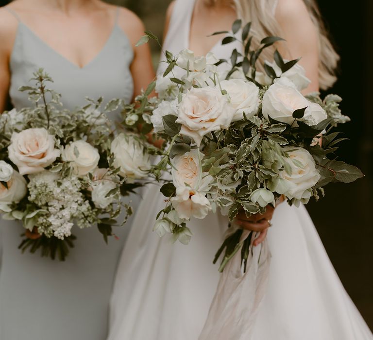 Bride & bridesmaid hold large white floral bouquets filled with roses and green foliage in classic styling