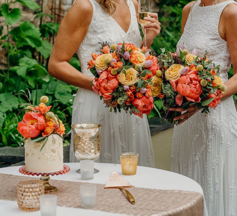 Brides hold bright and bold floral bouquets and glass of champagne next to small wedding cake with floral decor