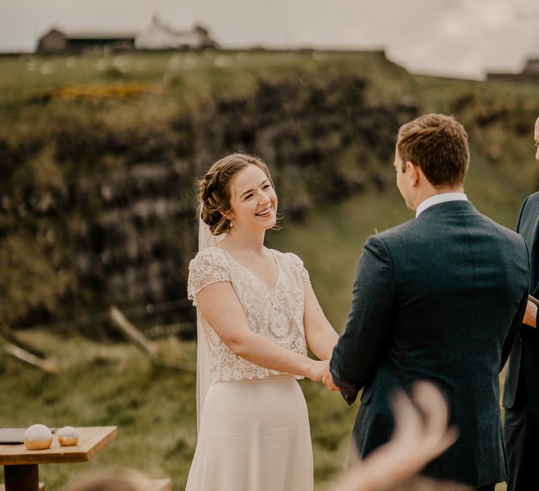 Smiling bride in lace top with capped sleeves and satin skirt holds hands with groom in navy suit at clifftop ceremony for Dunluce Castle wedding