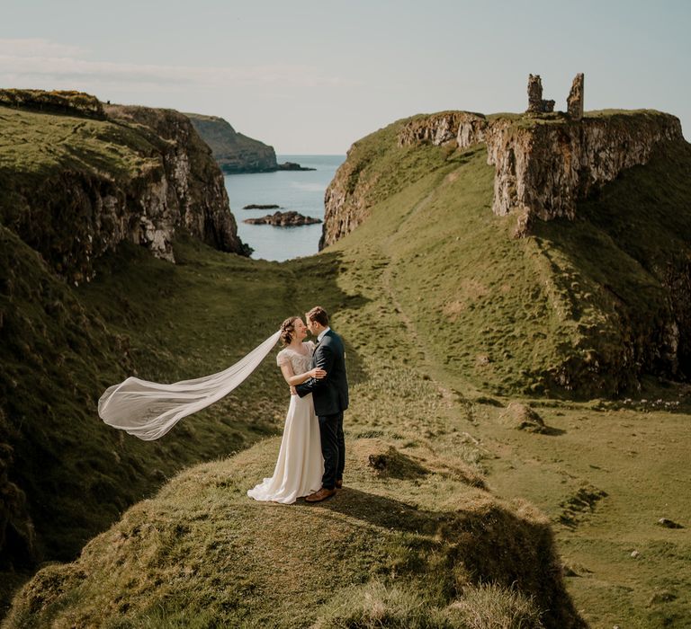 Bride in lace top wedding dress with satin skirt and veil blowing in the wind holds hands with groom on clifftop after Dunluce Castle wedding