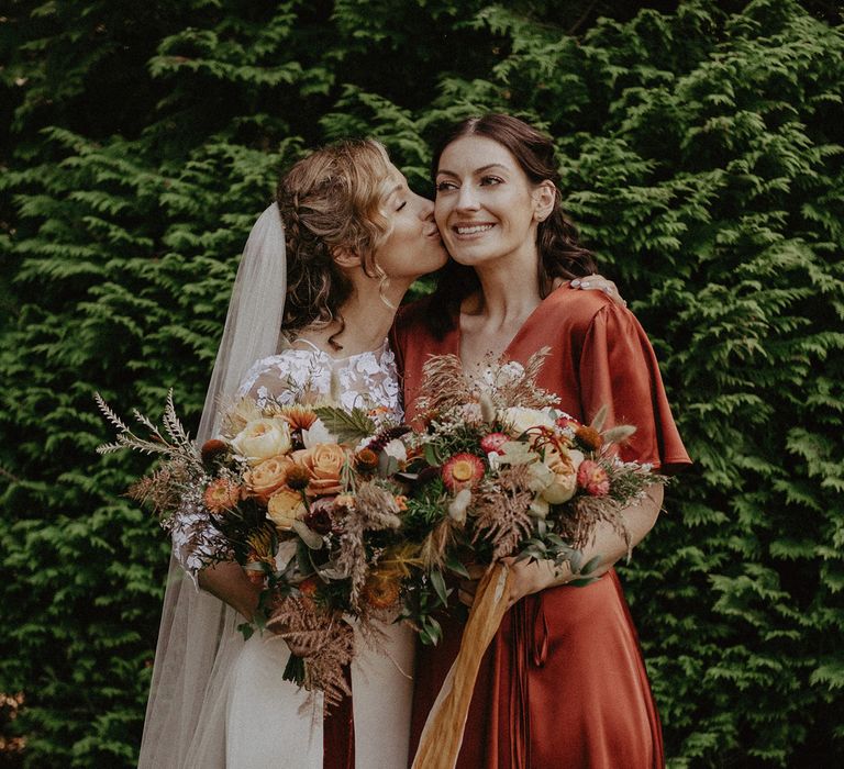 Bride in white Rime Arodaky wedding dress, mesh embroidered top and veil kisses cheek of bridesmaids in red satin bridesmaid dress whilst both holding burnt orange theme wedding bouquets at garden wedding