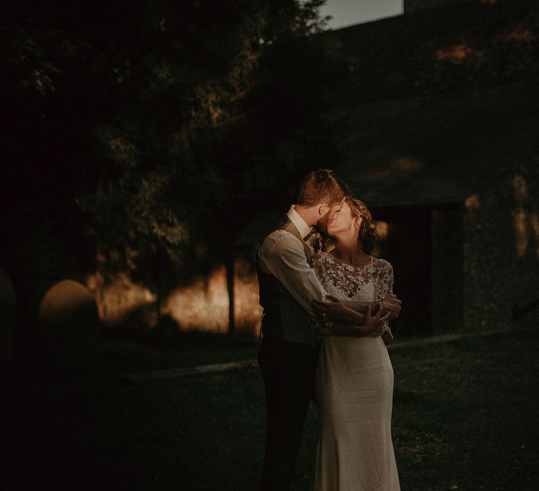 Bride in white Rime Arodaky wedding dress and mesh floral embroidered top kisses groom in white shirt and waistcoat in churchyard during golden hour at garden wedding with burnt orange wedding theme