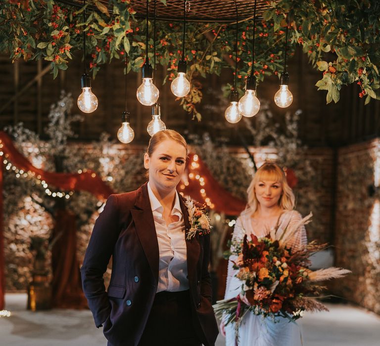 Bride in a plum coloured suit, with her wife looking on adoringly behind her