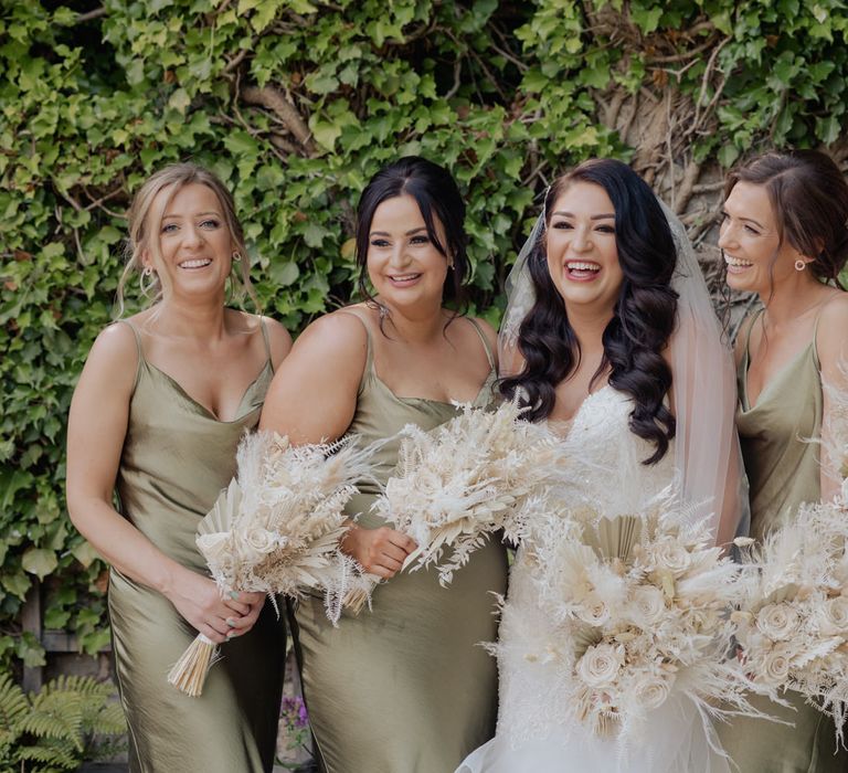 Bridesmaids in sage green satin dresses holding neutral dried flower wedding bouquets standing next to the bride 