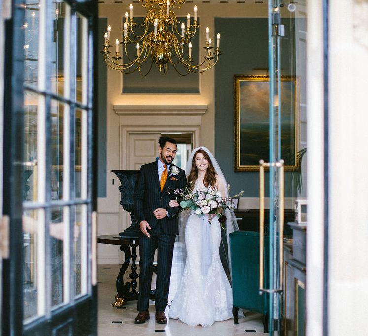 Grooms in a black suit and orange tie arm in arm with his bride in a lace Pronovias wedding dress at Grantley Hall wedding venue 