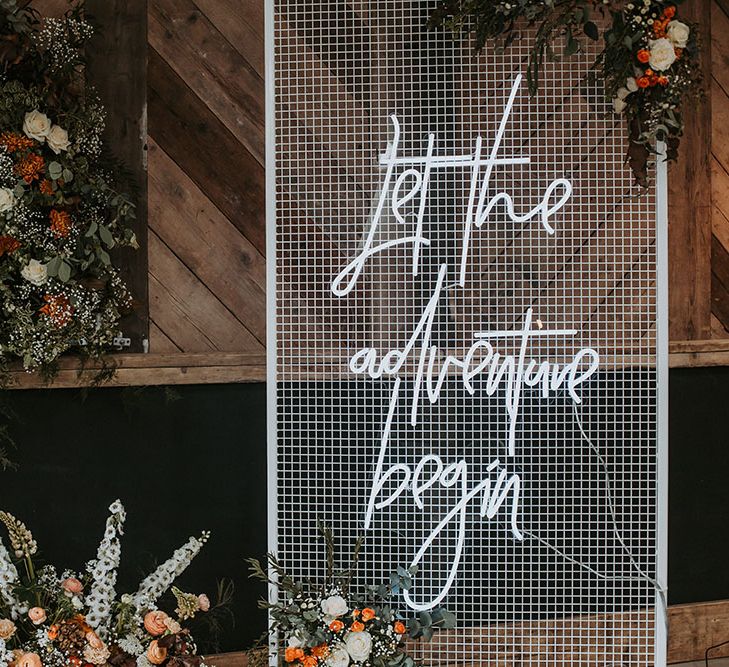 Let the adventure begin white neon wedding sign on a wire stand with orange, white and green wedding flowers 