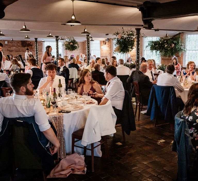 Wedding guests sit at round tables with white tablecloths and rose gold sparkle table runners in industrial wedding reception room with pendant lighting, exposed brick and fairy lights at The West Mill Derby