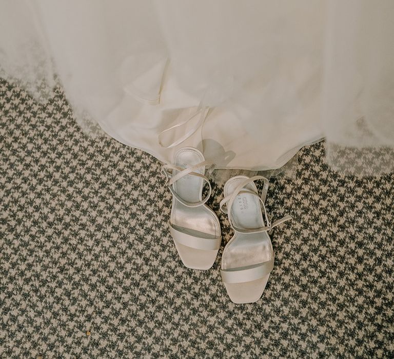 White strappy bridal shoes sit below wedding gown on the morning of wedding day