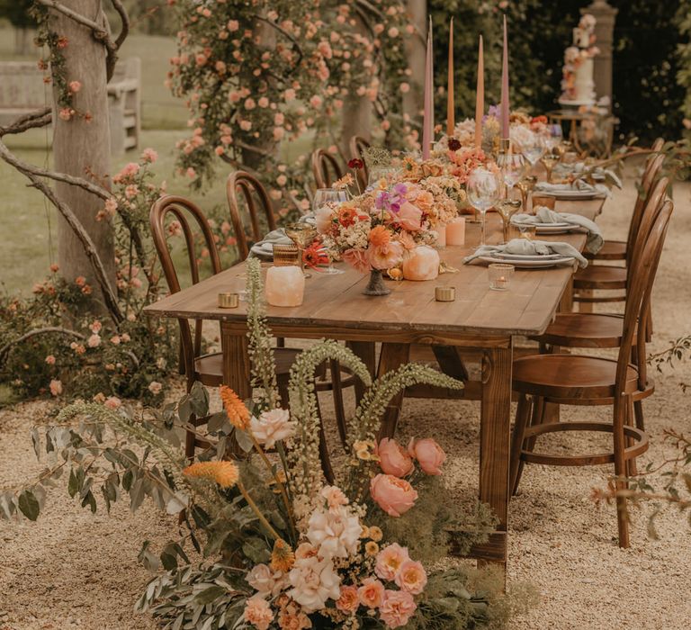 Outdoor wedding reception under a pergola with wooden table, pastel flowers, gold candlesticks and iridescent glassware 