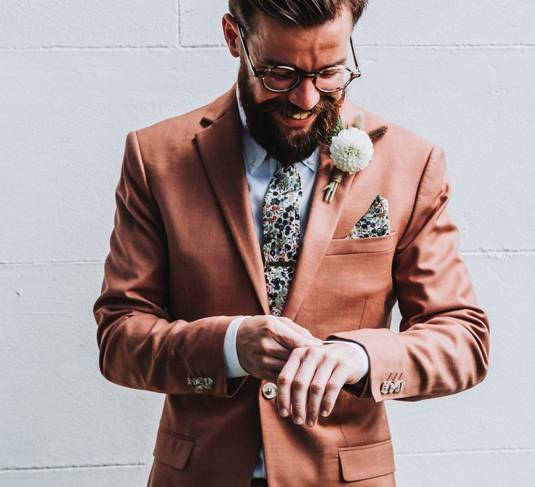 Groom adjusts his shirt sleeve on wedding day as he wears coral coloured suit complete with colourful tie and floral buttonhole 