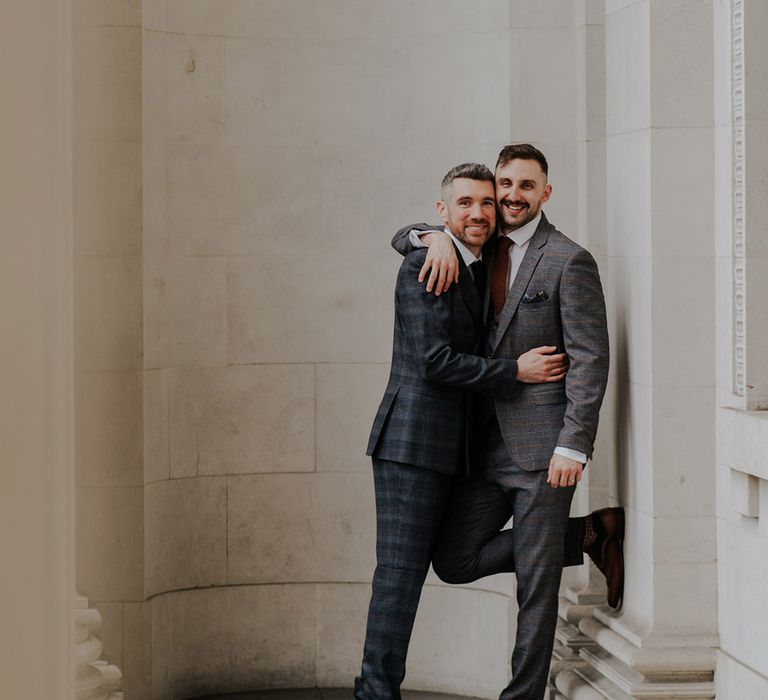 Grooms embrace one another in front of white walls on their wedding day for last minute wedding