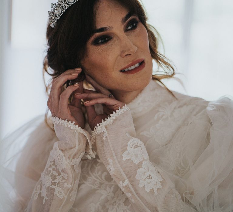 Bride applies her earrings as she wears long sleeved lace wedding gown with high neck