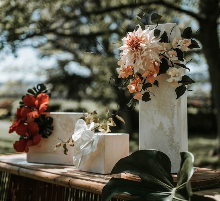 Three rectangular white marble iced cakes with tropical flower decor sit on rattan sideboard outdoors for birds of paradise wedding inspiration