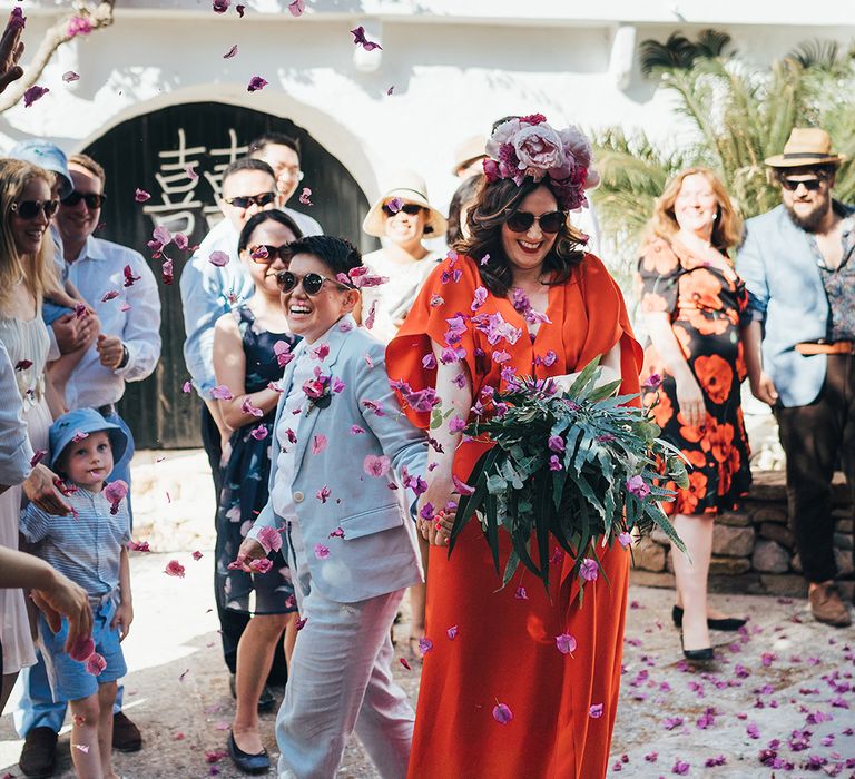 A lesbian couple hold hands as they walk through confetti. A Chinese bride wears a blue suit and white bride wears a red dress and flower crown.