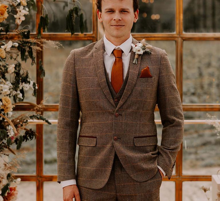 Stylish groom in a brown check wedding suit with rust coloured tie and pocket square and white buttonhole flower wrapped in twine 