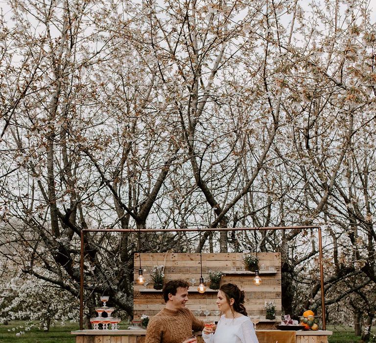 Bride in a laser cut lace wedding skirt and cashmere jumper having a cocktail at the wooden bar with her husband in chinos and a cable knit jumper