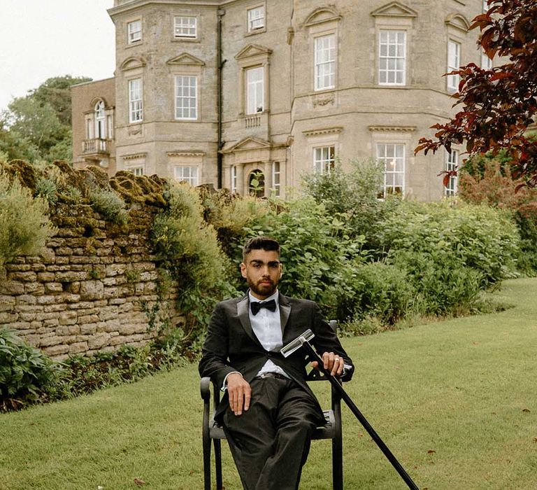 Stylish interabled groom in a black tuxedo sitting on a chair in the gardens at Bourton Hall with his walking stick 