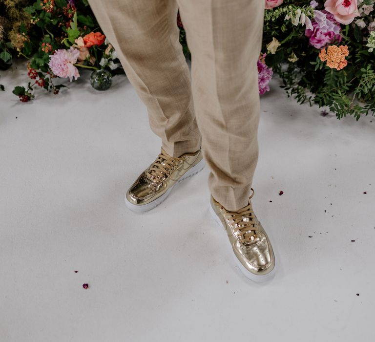 Groom in linen suit trousers stands by floral installation wearing gold Nike Air Force 1 trainers for wedding at Loft Studios London