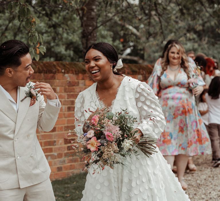 Laughing bride in white long mesh sleeve spotted Vagabond wedding dress and bridal hair bow holding mixed bridal bouquet stands with groom in shawl collared shirt and double breasted linen suit after guests have thrown confetti at Wasing Park wedding