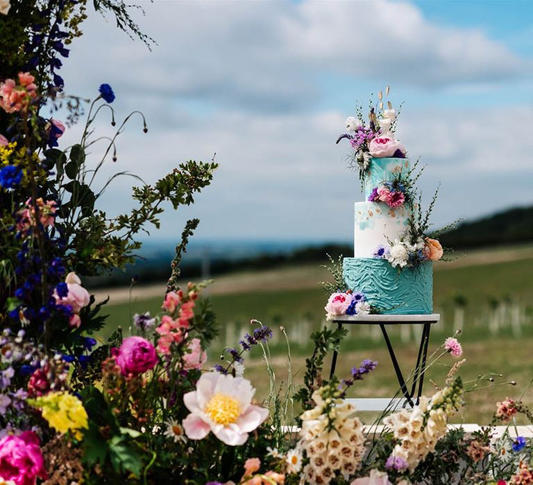 Three-tier wedding cake on a metal cake stand with turquoise and white buttercream layers decorated with colourful wild wedding flowers 