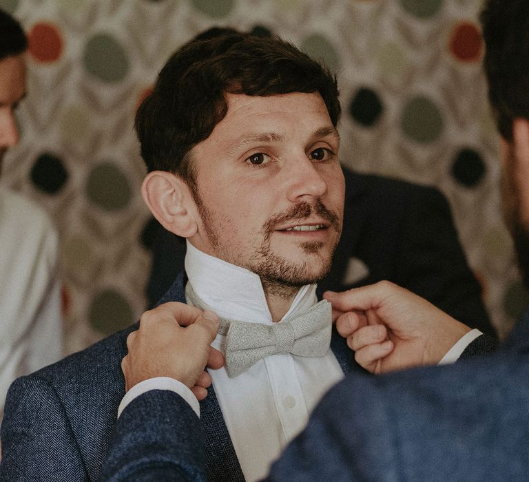 Groom in blue suit jacket and white shirt has his sage green bowtie adjusted by groomsman in blue suit jacket before Isle of Wight wedding with macrame wedding decor