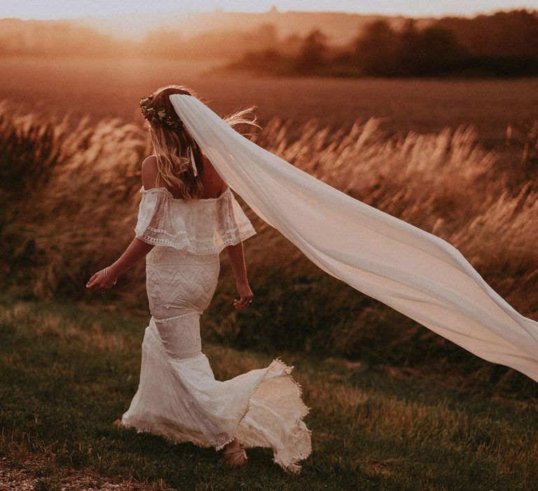 Bride in Grace Loves Lace bardot wedding dress, veil and flower crown walks by field at golden hour with groom in white shirt at Isle of Wight wedding with macrame wedding decor