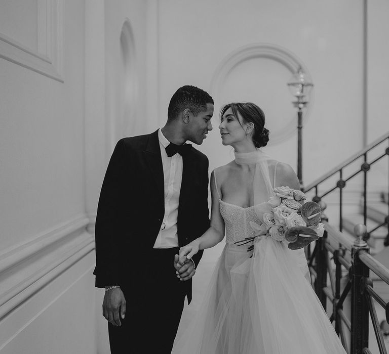 Black and white portrait of the bride and groom holding hands on the staircase of Clerkenwell House with the groom in a tuxedo and the bride in a romantic wedding dress with tulle skirt and sequin bodice 