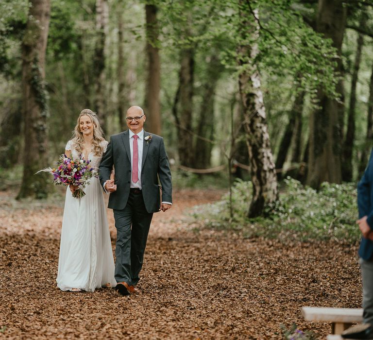 Bride in lace top wedding dress holding mixed bridal bouquet walks holding hands with father of the bride in dark suit and pink tie at woodland wedding ceremony in Norfolk