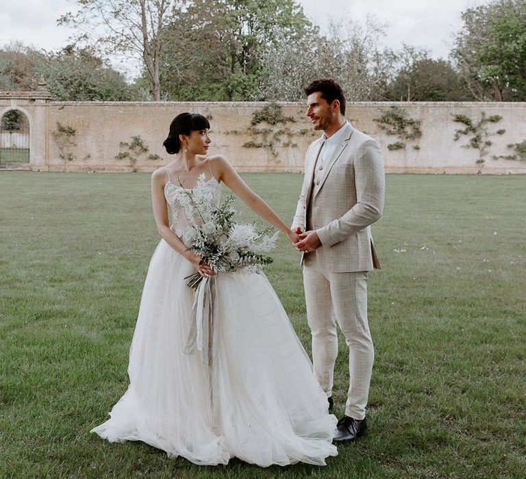 Groom in a slim fit three-piece beige check suit holding hands with his South Asian bride in a princess wedding dress