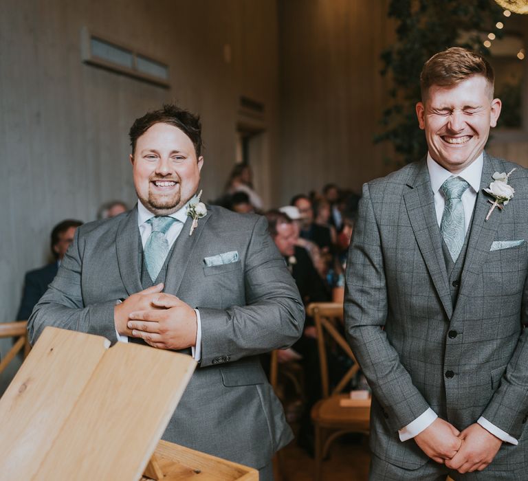 Groom in grey checked three piece suit stands with best man in grey suit both in mint green paisley ties and pockets squares as they wait for bride during wedding ceremony at Primrose Hill Farm