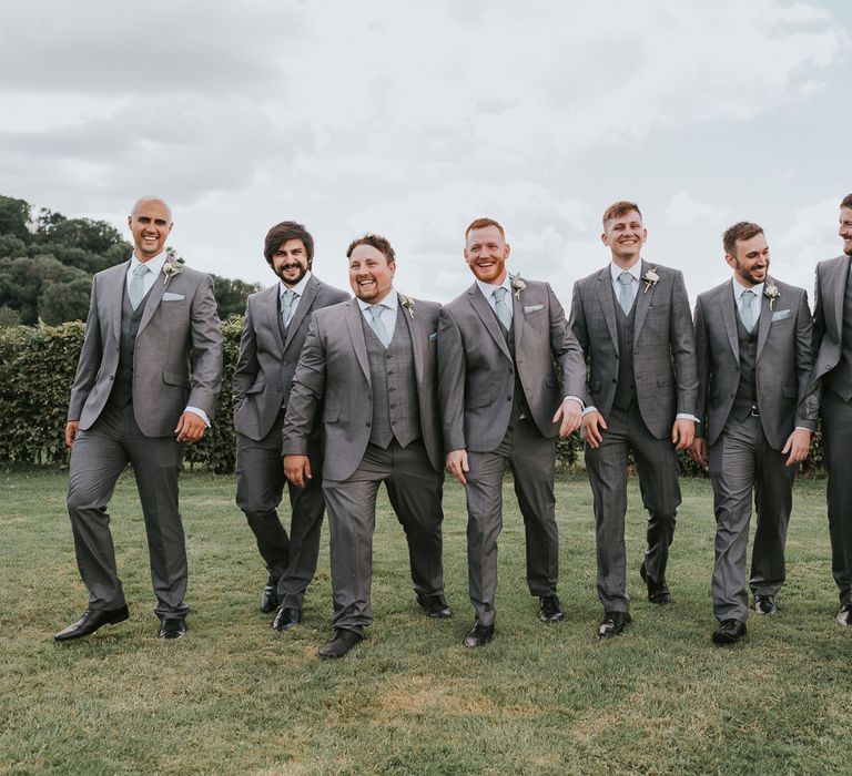 Groom and groomsmen in grey three piece suits with mint green paisley ties and pocket squares and floral buttonholes walk across grass during summer wedding at Primrose Hill Farm