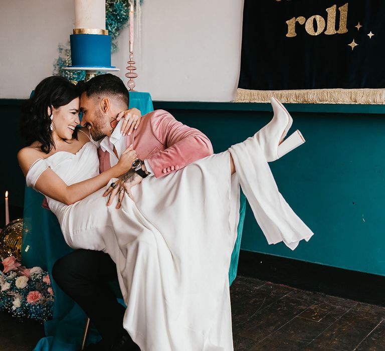 Bride in white wedding boots sitting on her grooms lap at their stylish pink and teal wedding reception 