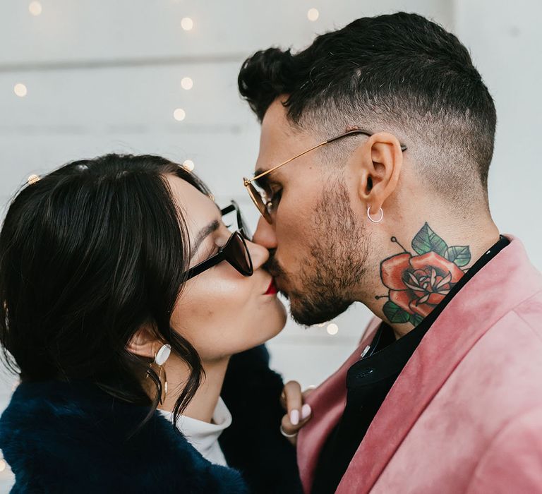 Groom in a pink velvet jacket kissing his bride in a navy blue faux fur coat and sunglasses 