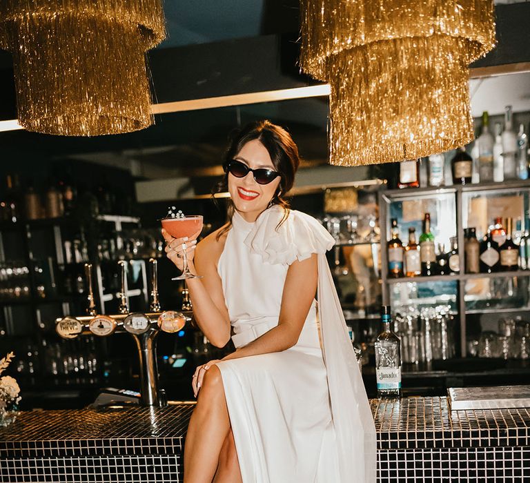 Bride in sunglasses with red lipstick sitting on the bar sipping cocktails in a wedding dress with front slit and white boots 