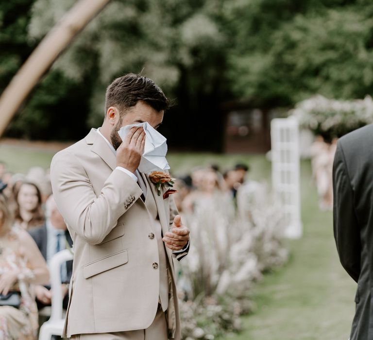 Groom in beige three piece Moss Bros. suit with floral buttonhole wipes eyes with white handkerchief whilst he waits for bride during DIY garden wedding ceremony