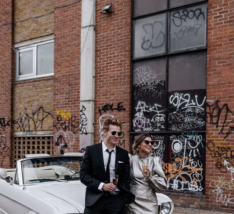 Cool urban bride and groom standing in front of vintage car sipping on bubbles Asylum London