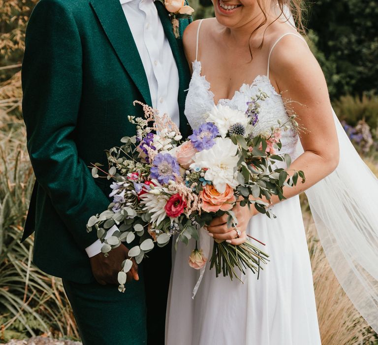 Black bride in a green wedding suit embracing her bride in a Grace Loves Lace wedding dress holding a colourful wedding bouquet