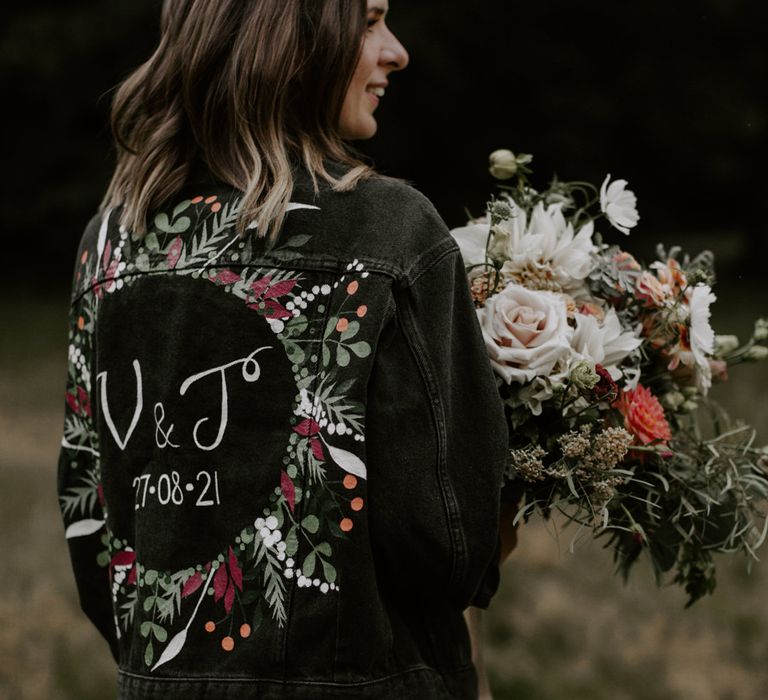 Bride wears personalised denim jacket with floral illustrations whilst holding her bouquet