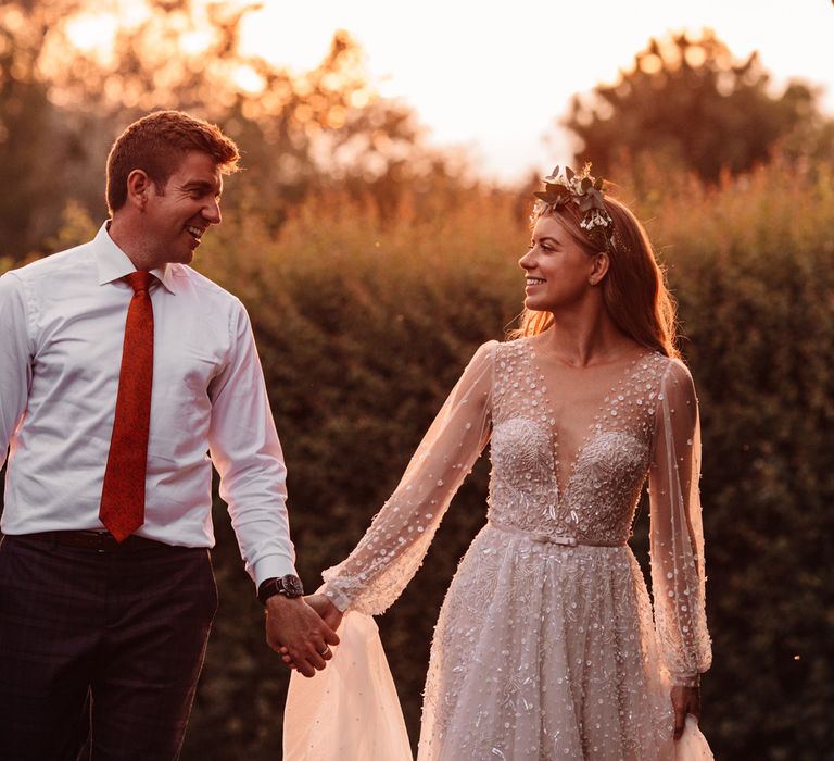 Bride in long mesh sleeve wedding dress with v neck and flower crown smiles whilst holding hands with groom in white shirt and red tie during golden hour