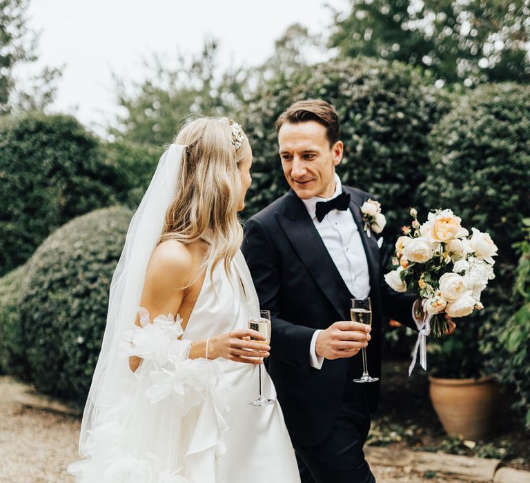 Bride in halterneck Halfpenny London wedding dress and Jimmy Choo wedding shoes walks with groom in black tie holding brides white and peach bouquet as they both hold champagne flutes