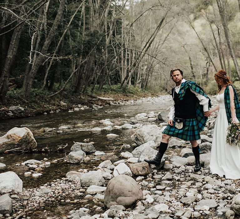 Groom in a tartan kilt and aran jumper holding hands with his bride in an a-line wedding dress as they cross a steam at Big Sur California 